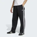 Pantalón deportivo Adidas Firebird de corte recto y holgado, color negro con las clásicas tres tiras blancas a lo largo de los laterales y el logo del trifolio bordado en la pierna izquierda.
