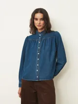 Camisa de jean de corte recto con cuello alto tipo mao con volados, cierre frontal con botones a presión y mangas largas con hombros ligeramente abullonados.