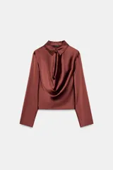 Blusa de tejido satinado color bordó, con cuello solapa, manga larga y detalle de abertura drapeada en el pecho. Cuenta con cierre frontal mediante un botón forrado.