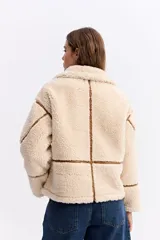 Campera tipo teddy de textura suave y acabado de peluche en color beige. Presenta cuello amplio con detalle de hebilla, cierre frontal y recortes en contraste de color marrón que marcan la estructura de la prenda.