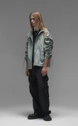 Campera rompevientos de diseño oversize, color verde, confeccionada en microfibra técnica ligera. Presenta capucha integrada, cierre frontal técnico y un sistema de frunces elásticos regulables a lo largo de las mangas que permiten ajustar el volumen de la prenda.