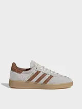 Zapatillas Adidas Handball Spezial de gamuza en tono gris claro, con las icónicas tres tiras laterales y refuerzo en el talón con textura de piel de serpiente en color marrón. Presenta suela de goma color caramelo y detalles dorados en el logo lateral y la lengüeta.