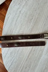 Cinturón de cuero vacuno color chocolate con apliques metálicos circulares grabados y tachas decorativas. Cuenta con hebilla metálica en tono bronce.