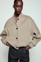 Cazadora bomber beige confeccionada en hilatura con lana, con cuello subido, manga larga y bolsillos frontales. Presenta cierre frontal con botones oscuros y puños, cuello y ruedo en tejido rib.