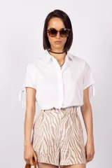 Blusa blanca de algodón con cuello camisero, manga corta con lazo ajustable y cierre frontal con botones.