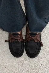 Mocasín de estilo náutico confeccionado en cuero, combinando una base de cuero con textura de pelo en color negro y detalles de cuero con textura croco en color marrón. Presenta cordones decorativos con borlas laterales y suela plana de goma.