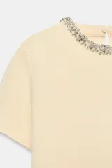 Jersey de punto color amarillo claro, con cuello redondo adornado con aplicación de joyas a contraste, manga corta y cierre en la espalda con abertura y botón.
