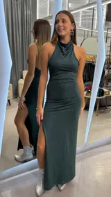 Vestido largo verde oscuro con acabado brillante, de corte ajustado, escote halter con cuello alto fruncido y tajo lateral en la pierna.