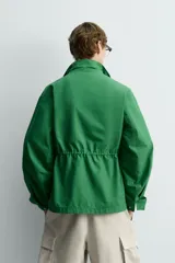 Chaqueta tipo cazadora de corte relajado en color verde, confeccionada en tejido de mezcla de algodón. Presenta cuello subido con forro interior a cuadros, manga larga con puños abotonados y cuatro bolsillos frontales de plastrón con solapa. Incluye cierre frontal de cremallera oculta por solapa con botones a presión y cintura ajustable en el interior.