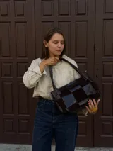 Cartera de hombro confeccionada en cuero y gamuza con diseño de damero, combinando paneles de texturas lisas y aterciopeladas. Presenta una manija trenzada y detalles de costuras artesanales en los laterales.