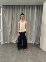 Pantalón de jean azul raw con diseño de corte versátil, ajustable mediante cierres laterales que revelan un panel interior rojo. Presenta una estampa gráfica de gran tamaño en la parte trasera con detalles bordados, costuras reforzadas y etiqueta de cuero en la cintura.