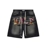 Short de jean estilo jorts con lavado desgastado en color negro. Presenta letras grandes bordadas con parches de tela a cuadros multicolor y detalles de tachas metálicas. Incluye cordones largos en la cintura y bordado adicional en la parte inferior.