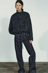 Cazadora de denim azul oscuro con cuello solapa, cierre frontal con cremallera y botones a presión ocultos. Presenta mangas largas tipo murciélago con puños, bolsillos de vivo en el delantero y bajo elástico ajustable con cordones.