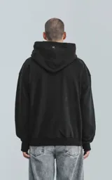 Hoodie negro de frisa premium, corte oversize, con capucha ajustable y puños y cintura elastizados. Presenta un bolsillo canguro frontal y un detalle de etiqueta tejida con texto repetido en los cordones de la capucha y en la manga izquierda.