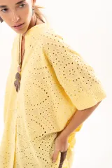 Camisa de corte oversize confeccionada en tela bordada con diseño calado, color amarillo. Presenta cuello mao, cierre frontal con botones y mangas cortas amplias.
