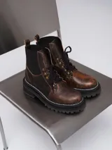 Borcego de gamuza color chocolate con paneles laterales elásticos negros y suela track dentada. Presenta sistema de cordones con ganchos metálicos y tirador en la parte trasera.