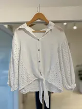 Camisa blanca de lino con mangas abullonadas y detalles de bordado inglés en las mangas y en los laterales. Presenta cuello camisero, cierre frontal con botones y tiritas para atar en el frente.