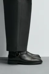 Bota de caña media fabricada en piel color negro, con detalle de arnés y hebilla metálica lateral. Presenta horma cuadrada y suela a tono con ligero tacón.