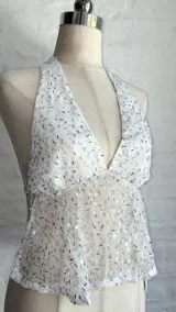 Top halter blanco con escote pronunciado en V, confeccionado en tejido transparente con lentejuelas o brillos plateados. La parte superior del busto es opaca y el resto del cuerpo es traslúcido.