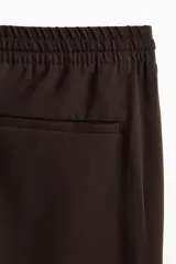 Pantalón relaxed fit color marrón, confeccionado en tejido suave, con cintura elástica ajustable mediante cordón, bolsillos laterales discretos, bolsillo posterior insertado y piernas rectas con pliegues frontales y traseros.