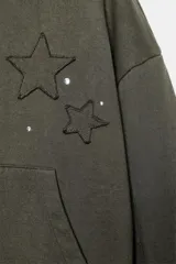 Sudadera con capucha y bolsillo tipo canguro, confeccionada en tejido con efecto lavado. Presenta diseño de estrellas bordadas y aplicación de tachas decorativas en el frente, con terminaciones en rib en puños y ruedo.