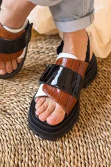 Sandalias de plataforma con suela track negra. Presentan un diseño de tiras anchas combinadas en cuero con textura de reptil color marrón y una franja central de charol negro. Incluyen una tira trasera elástica para mayor sujeción.