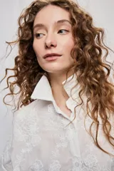 Vestido camisero corto y de silueta amplia en color blanco, con cuello clásico, cierre frontal de botones y mangas largas. Cuenta con diseño bordado floral en toda la prenda y dobladillo asimétrico, más largo en la parte trasera.