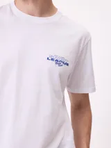 Remera de manga corta color blanco con estampa gráfica en la espalda en color azul, que presenta un diseño de estilo surf con una camioneta, un pelícano y texto alusivo a la marca Rusty.