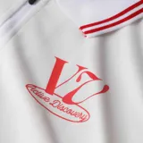 Chomba blanca con cuello tipo polo con cierre de cremallera corto y ribete rojo. Tiene un logo estampado en el pecho.