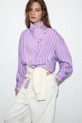 Camisa de corte oversize con diseño de rayas verticales en tonos violeta, blanco y bordó. Presenta cuello subido cruzado con cierre de botones frontal, mangas largas con puños y bajo asimétrico.