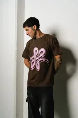 Remera de manga corta, corte regular fit, color marrón oscuro con un gran estampado frontal en rosa pálido que simula un logo estilizado o un símbolo abstracto.