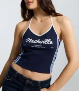 Top frente única de algodón en color azul marino con ribetes blancos en el cuello y rayas laterales. Presenta un estampado frontal con la inscripción Nashville y detalles en blanco. Diseño sin mangas con lazo para anudar al cuello.