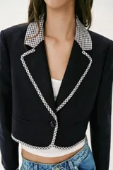 Blazer corta de color negro con cuello solapa, manga larga y hombreras. Presenta detalles combinados con estampado de cuadro vichy en el cuello, los bordes de la solapa, el ruedo y los puños. Cuenta con cierre frontal de un botón y falsos bolsillos delanteros de vivo.