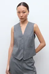 Chaleco sastrero de corte entallado con escote en V, cierre frontal con botones y bolsillos laterales simulados. Presenta un estampado de microcuadros en tonos grises.