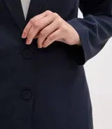 Blazer de corte sastre confeccionado en crepé, con diseño alargado y cierre frontal de doble abotonado con botones forrados. Presenta solapas clásicas y estructura recta.