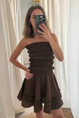 Vestido corto color marrón oscuro, sin mangas y con escote recto. Presenta un cuerpo fruncido con elástico y una falda corta con volados en el ruedo.