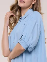 Camisa de tencel liviano estilo denim, color celeste, con cuello camisero, cierre frontal con botones nacarados, mangas cortas abullonadas con puños elásticos en punto smock y detalles de ojalillos.