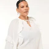 Blusa blanca de manga larga con textura de plumeti (pequeños puntos en relieve) y volantes decorativos verticales en el frente y en los puños. Presenta un escote en V con lazos para anudar en el cuello.