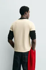 Camiseta de corte regular, color crema, con cuello redondo y ribetes en las mangas y cuello en color negro. Presenta un estampado frontal con un corazón rojo y texto en negro que dice "Everything beats for something".