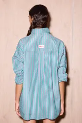 Camisa holgada de algodón con rayas verticales en tonos verde agua, rosa pálido y blanco. Presenta una abertura frontal que llega hasta la mitad del torso, ribeteada con un cordón ondulado de color verde brillante. Incluye botones oscuros y un pañuelo a juego en la cabeza.