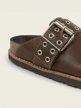 Zueco de cuero color chocolate, con diseño de punta redondeada y tira ajustable sobre el empeine con hebilla metálica y detalle de ojales decorativos.