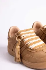 Zapatillas urbanas color tostado con textura geométrica en el cuerpo y detalles de gamuza en la puntera y laterales. Presentan un diseño distintivo con cordones anchos tejidos en tonos tostado y blanco, y un accesorio metálico con borla de gamuza en el empeine.