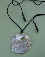 Collar artesanal con dije central de metal con textura orgánica irregular que evoca una ostra. El dije está montado sobre un cordón de algodón natural, permitiendo un uso versátil y regulable.