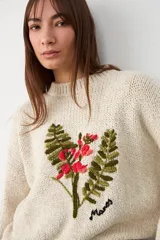 Sweater de punto color crudo, con cuello redondo y diseño de bordado floral en el frente. Presenta mangas largas con puños acanalados y un corte holgado.