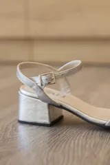 Sandalias de fiesta con diseño elegante y minimalista, confeccionadas en cuero con acabado metalizado plateado. Presentan una tira ancha sobre el empeine, puntera cuadrada, correa fina con hebilla ajustable al tobillo y tacón ancho de altura media.