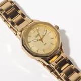 Reloj de pulsera dorado con malla metálica, caja octogonal y esfera dorada con agujas y marcadores de hora minimalistas.