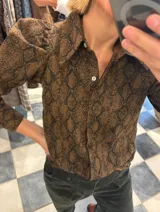 Camisa marrón con estampado de piel de serpiente, cuello clásico, mangas largas abullonadas y cierre frontal con botones.
