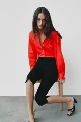 Camisa de popelina roja, de corte oversize, con cuello solapa, manga larga y bajo asimétrico redondeado. Cierre frontal oculto con botones.