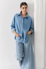 Camisa de jean azul claro con bordados pequeños en color blanco de figuras deportivas. Presenta cuello clásico, cierre frontal con botones de madera y mangas largas con puños abotonados.