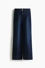 Pantalón de jean estilo wide leg con tiro alto, confeccionado en denim de algodón con leve stretch. Presenta cierre frontal con doble botón metálico, pliegues frontales y traseros, y bolsillos traseros de parche.